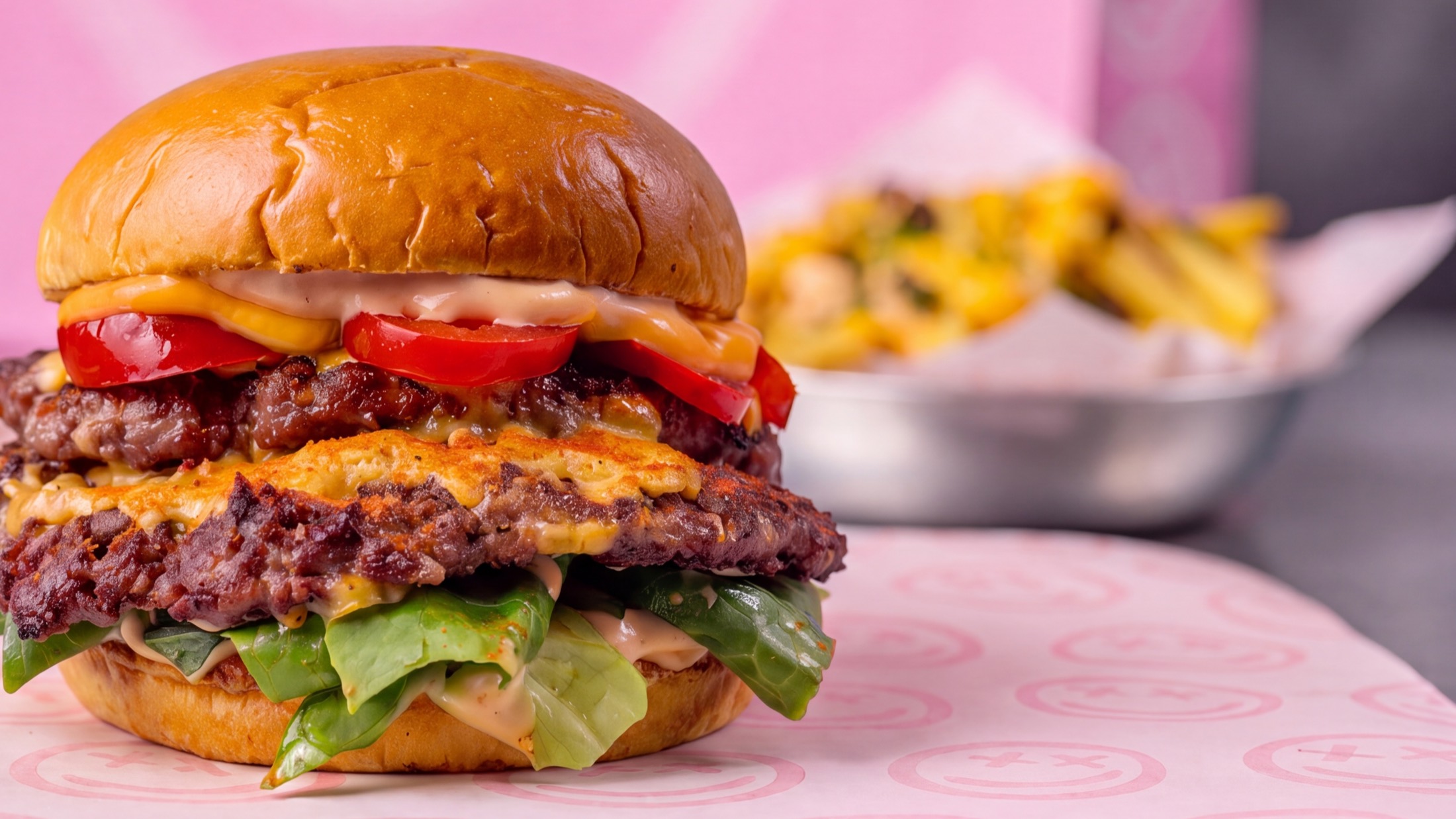 A double smashed burger with melted cheese, tomato and lettuce on greaseproof paper, with a pink Super Awesome Deluxe bag behind it.